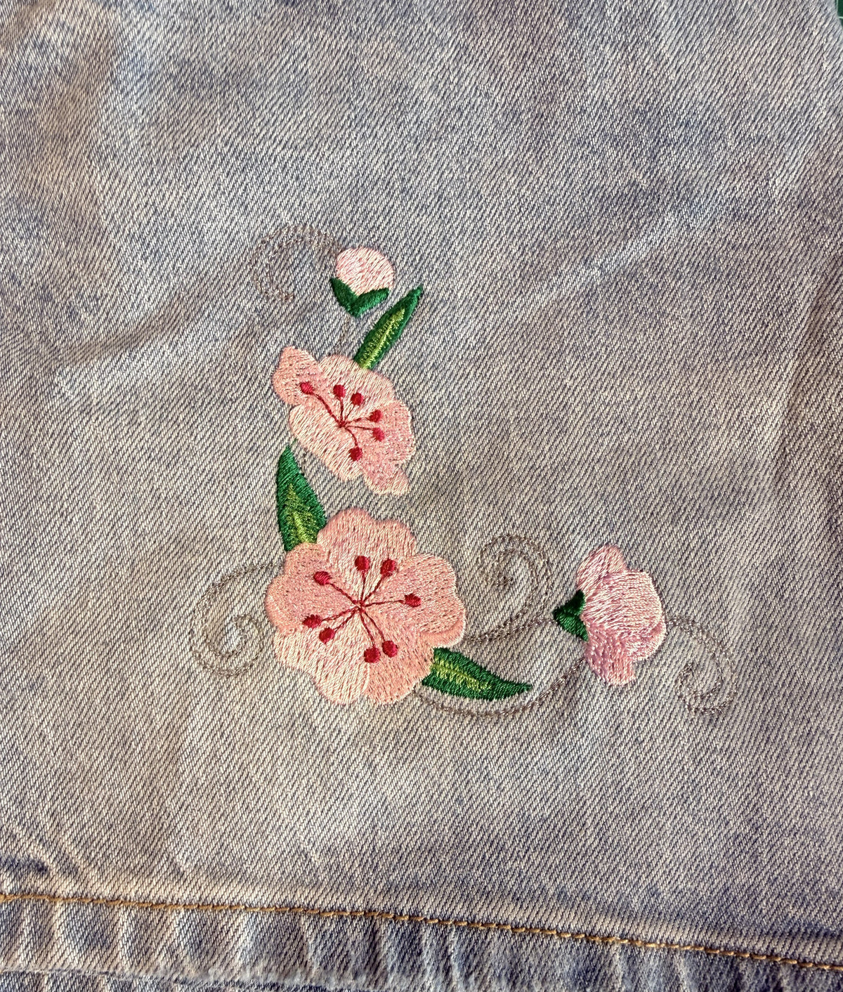A close-up of light blue denim fabric featuring a pink cherry blossom floral embroidery design with green leaves, placed on a green cutting mat.