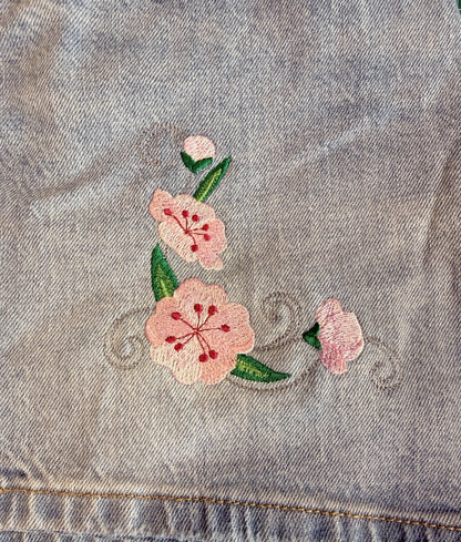 A close-up of light blue denim fabric featuring a pink cherry blossom floral embroidery design with green leaves, placed on a green cutting mat.